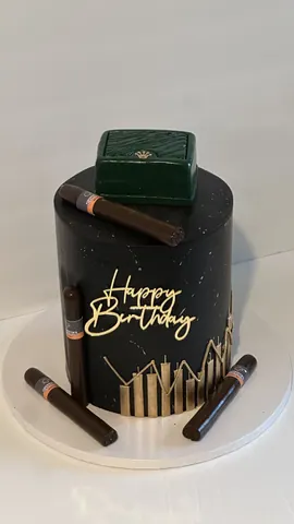 Tall black cake with a gold city skyline, a gold Happy Birthday sign, chocolate cigars and a green box on top.
