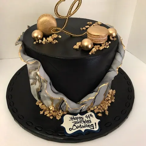 Black cake topped with a gold script letter, gold macarons, gold spheres and gold flakes.