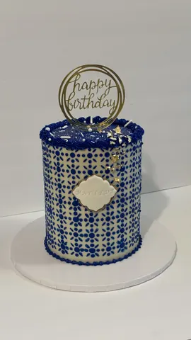 White cake covered in a blue tile pattern with a plaque and a gold ring Happy Birthday topper.