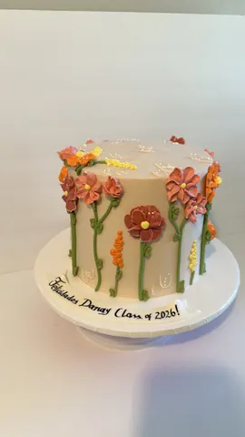 Cream cake covered in hand-piped buttercream wildflowers in orange, red and coral with green stems, for a graduation.