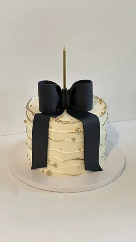 Cream cake with wavy gold-lined ridges, small gold pearls, a large black bow and a tall gold candle.