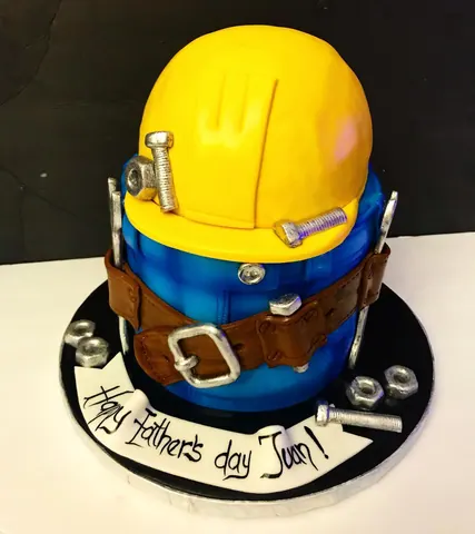 Sculpted cake of a yellow construction hard hat on a blue work shirt with a brown tool belt, sugar bolts, nuts and a wrench, for Father’s Day.