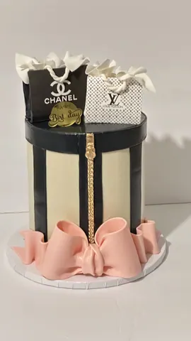 Hat box cake in black and cream with a gold zipper, pink bows around the base and small shopping bags on top.