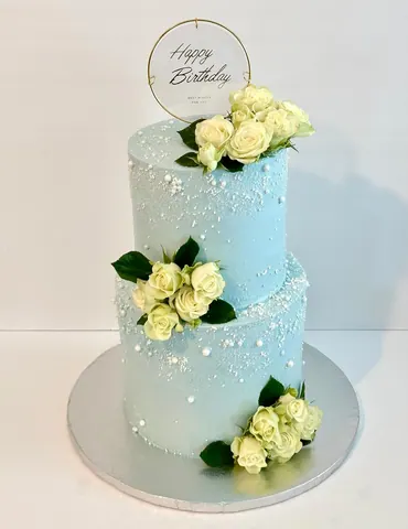 Two-tier light blue cake with clusters of white roses, scattered pearls and a gold ring Happy Birthday topper.