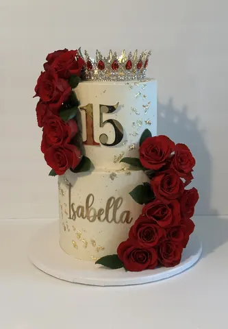 Two-tier ivory cake for a 15th birthday with a cascade of red roses, a jeweled gold crown, a gold 15 and a gold script name.