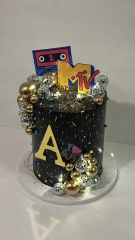 Black retro party cake with a gold letter A, gold and silver disco balls, confetti sprinkles and a cassette tape topper.