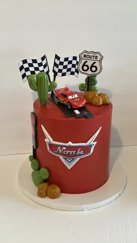 Red birthday cake with a race car on a road, cactus, a route sign, checkered flags and a name badge.