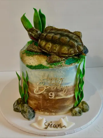 Cake sculpted as a realistic bronze tortoise resting on green grass, with tall green leaves behind it.