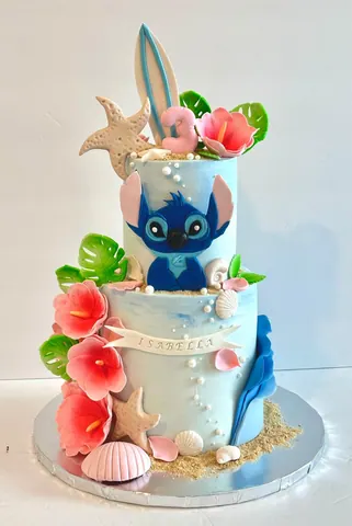 Two-tier beach cake in sea blue and sand with pink hibiscus flowers, palm leaves, sea shells, a starfish and a surfboard topper.