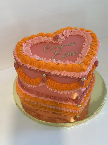 Heart-shaped vintage cake in pink with ruffled orange and pink buttercream piping, gold pearls and a script message on top.